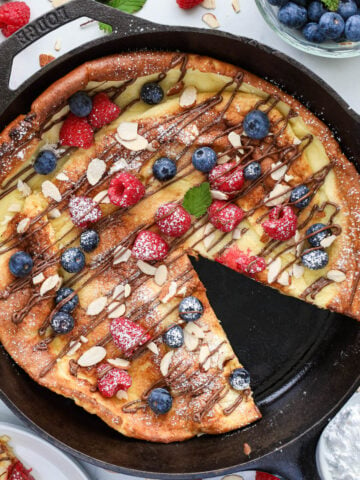 German Pancake in a cast iron skillet topped with Nutella, sliced almonds, berries, and powdered sugar, one slice missing.