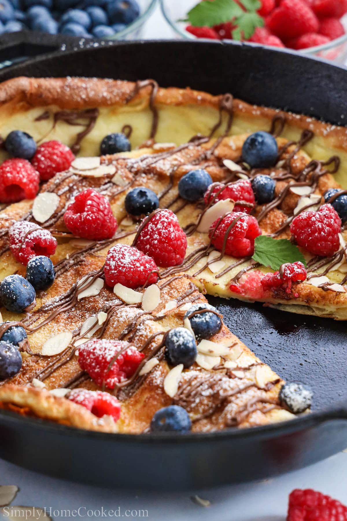 German Pancake in a cast iron skillet missing a piece and topped with almonds, powdered sugar, berries, and nutella.