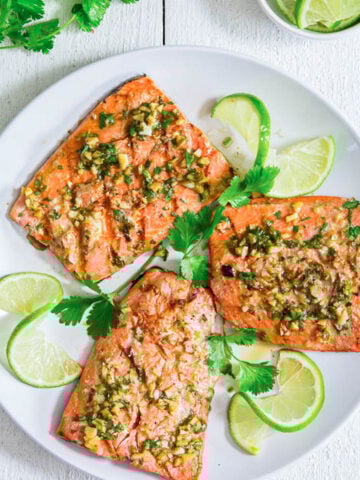 Cilantro Lime Salmon covered in marinade and on a white plate with lime slices and cilantro.
