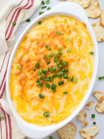 Crab Artichoke Dip in a serving dish topped with cheese and green onion with crackers nearby.