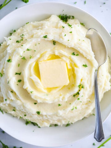 Bowl of Super Creamy Mashed Potatoes with a spoon and topped with a pat of butter and parsley.