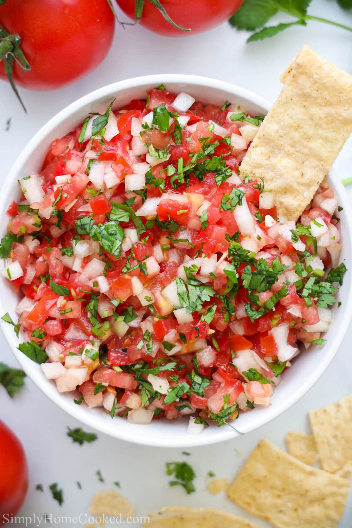 Overhead view of a white bowl of Pico de Gallo with a chip in it and more nearby