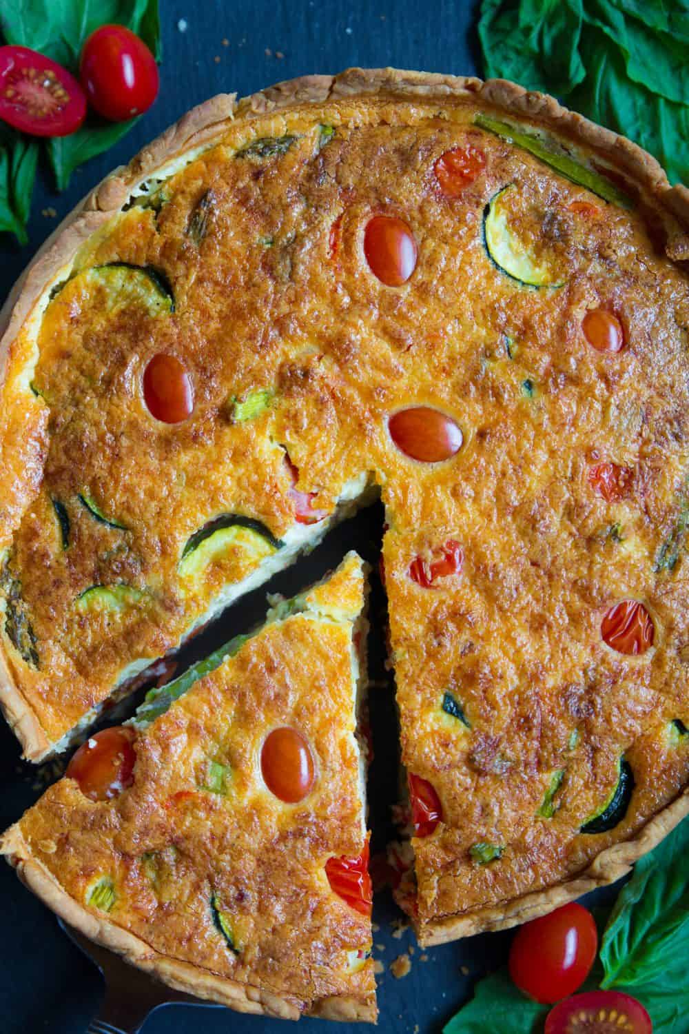 An overhead image of a baked Quiche Lorraine with zucchini, cherry tomatoes, and asparagus, with basil leaves beside it.