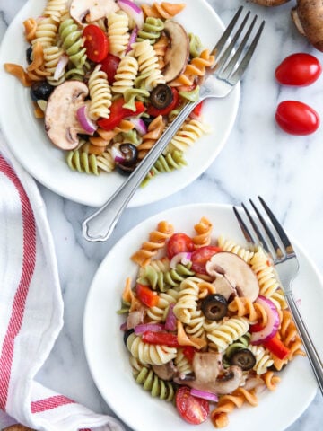 Two plates of Tri Color Pasta Salad with forks and mushrooms and cherry tomatoes nearby.