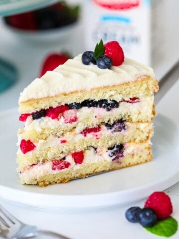 sliced Chantilly cake with berries on top on a white plate with a blue cake stand behind it