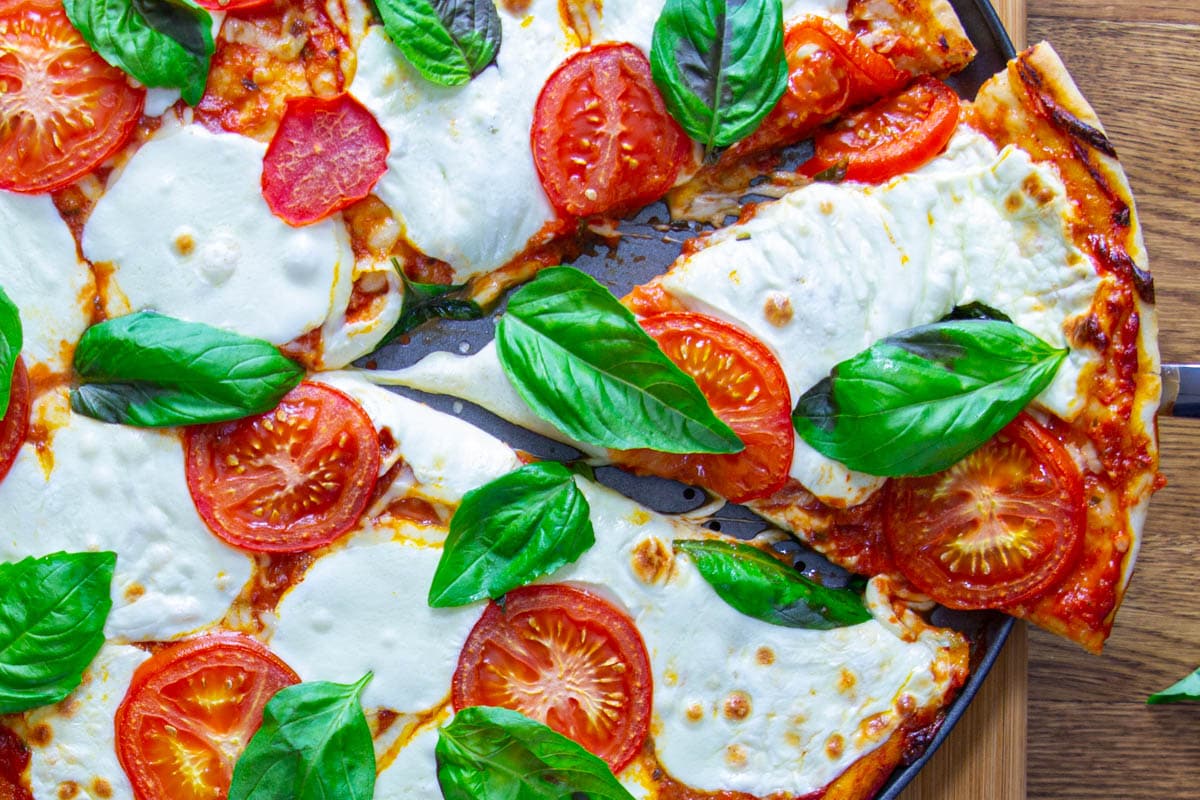 A slice being removed from a Margherita Pizza topped with tomatoes, mozzarella, and basil leaves.