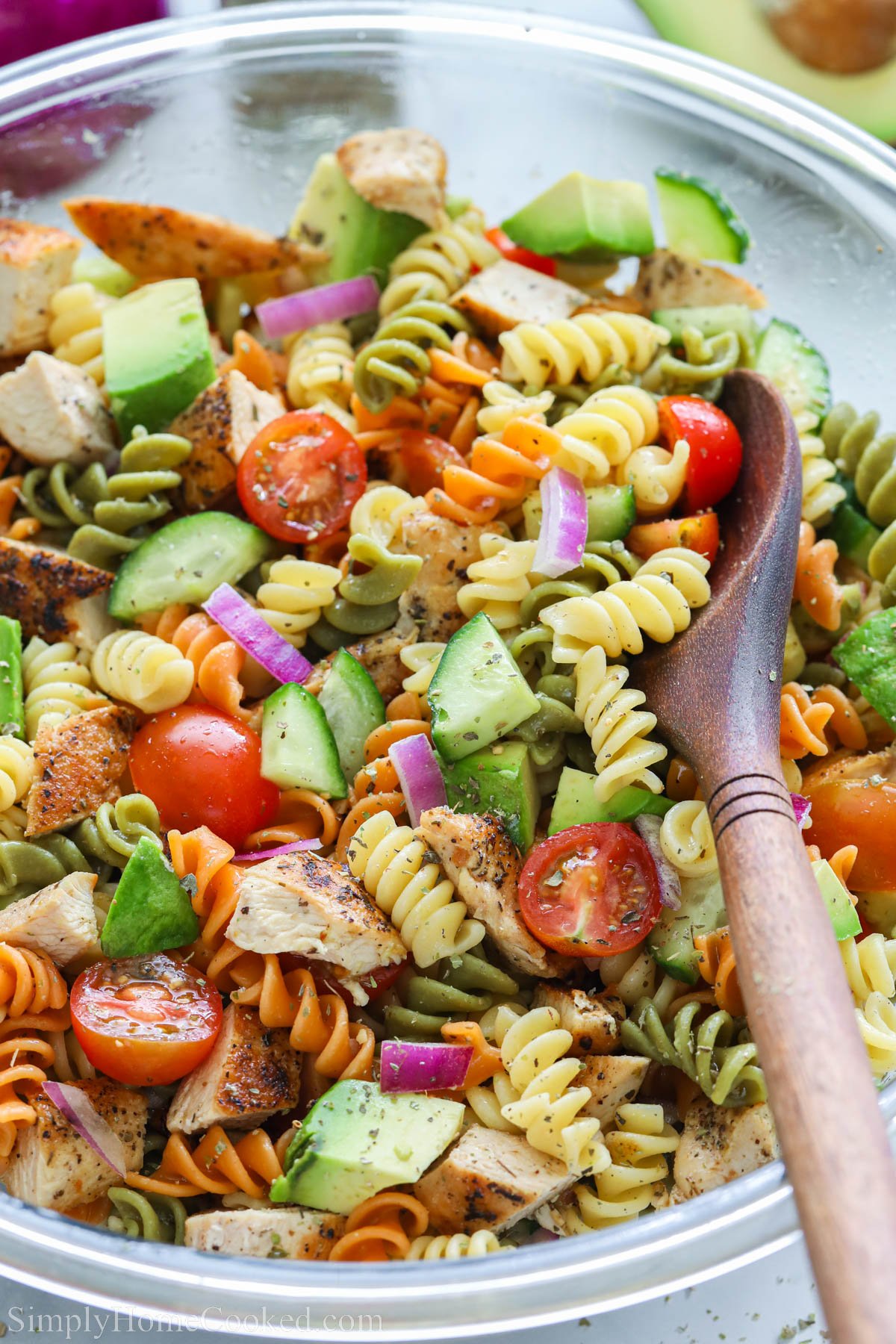 Chicken Pasta Salad with tomatoes, cucumber, avocado, onions, rotini, and chicken in a bowl with a wooden spoon.
