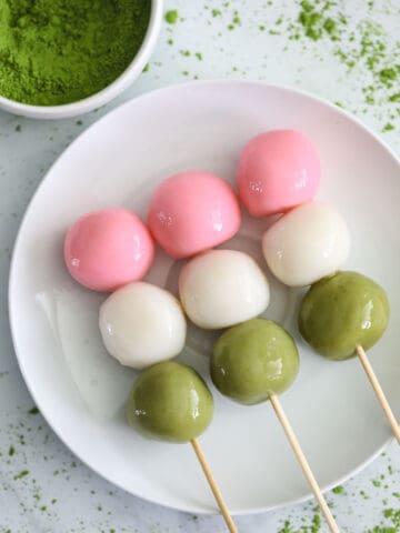 3 Hanami Dango on skewers on a white plate with matcha powder nearby.