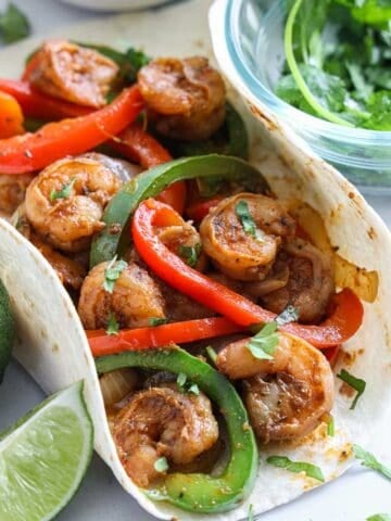 Vertical close up of Shrimp Fajitas in a flour tortilla, cilantro and a lime wedge on the side.