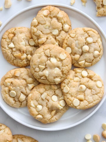 Vertical image of a plate of White Chocolate Macadamia Nut Cookies with nuts and chocolate chips nearby