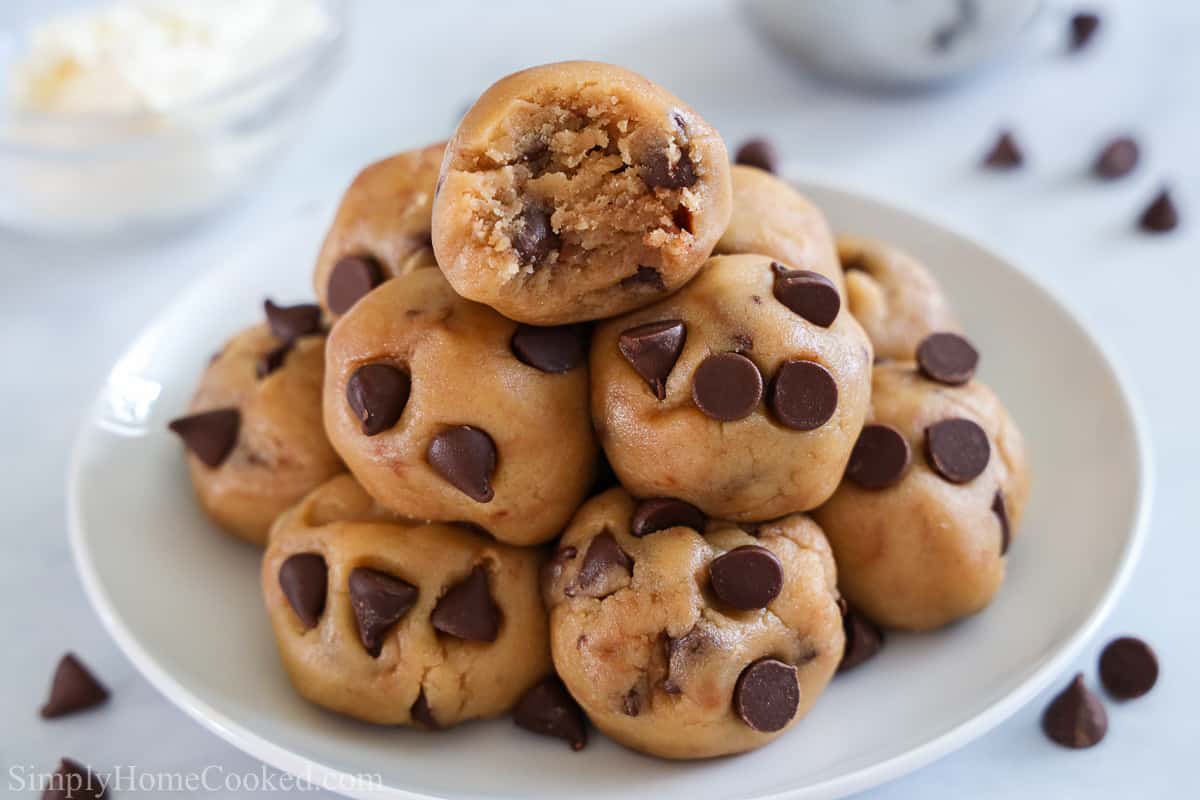 Balls of Quick Edible Cookie Dough in a pile on a white plate with one missing a bite and scattered chocolate chips in the background.