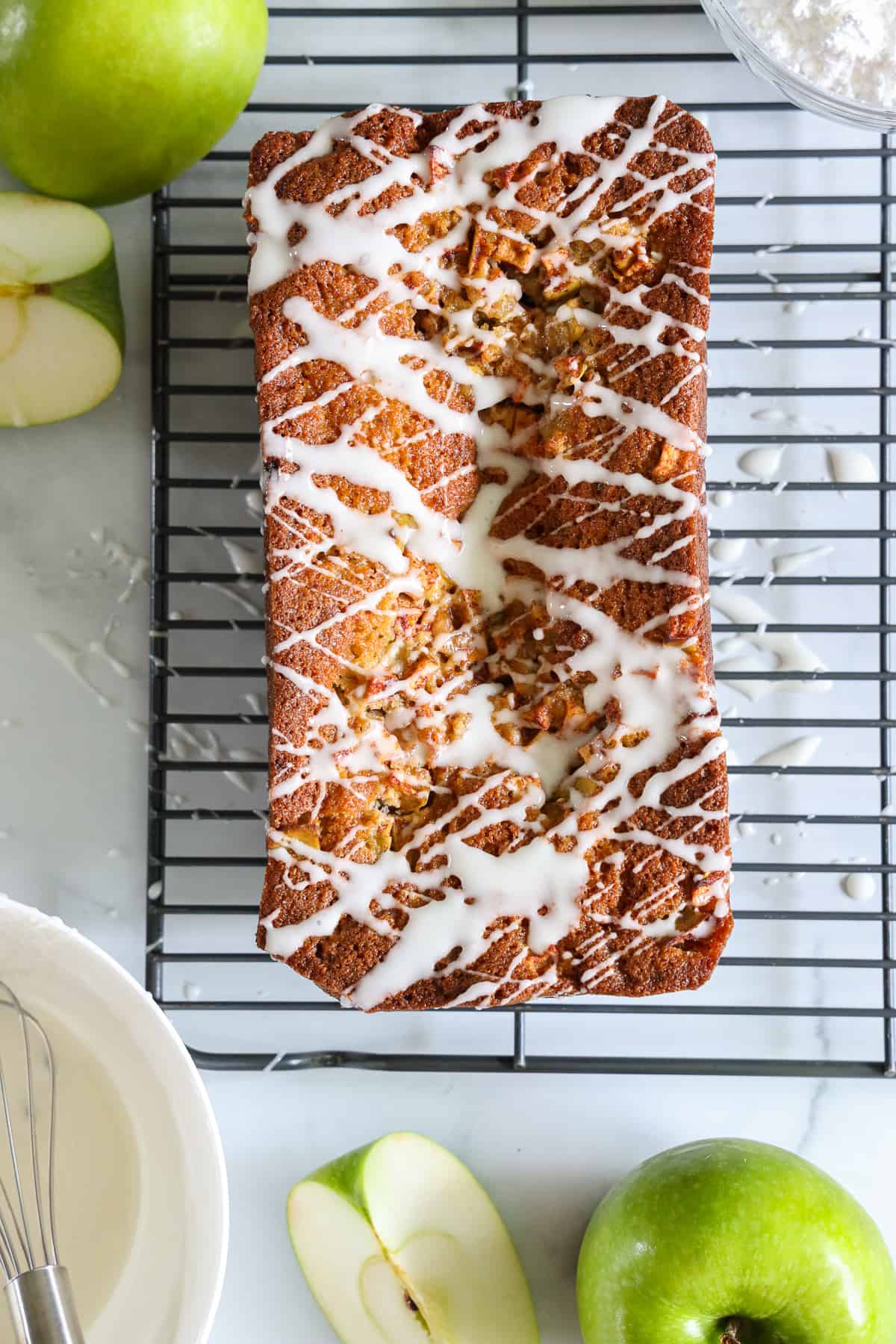 Overhead view of Apple Fritter Bread, with glaze drizzled on top as it sits on a cooling rack, with green apples to the side.