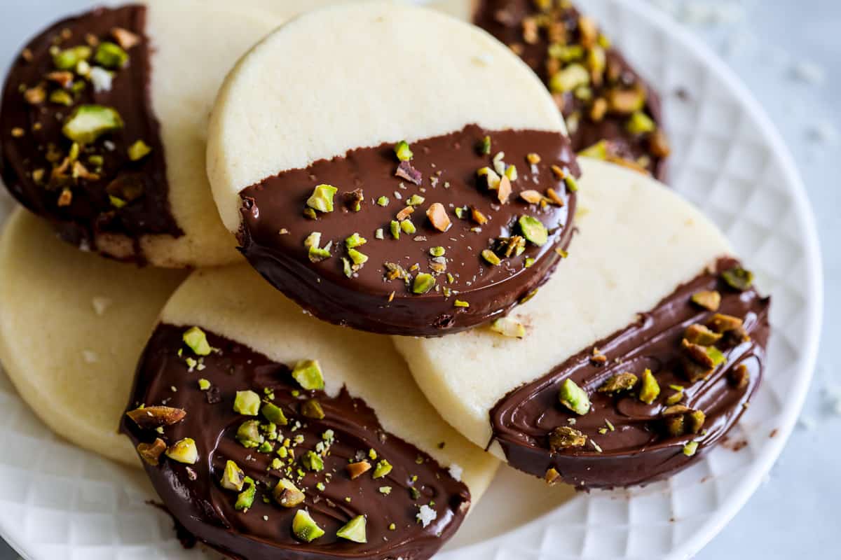 Classic Shortbread Cookies half dipped in chocolate with chopped pistachios on top, sitting in a pile on a white plate.