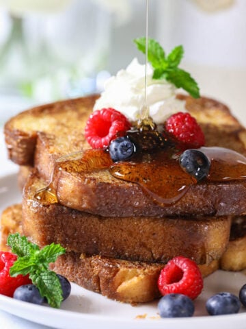 Syrup drizzling on a stack of Brioche French Toast on a plate with fresh berries and whipped cream