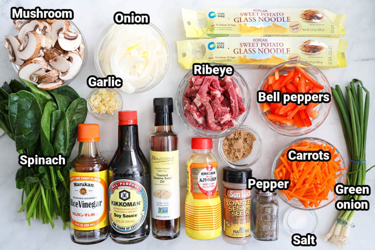 Ingredients in Easy Japchae Recipe: mushrooms, onion, garlic, spinach, glass noodles, ribeye, bell peppers, carrots, green onions, salt, pepper, brown sugar, toasted sesame seeds, aji mirin, toasted sesame oil, soy sauce, and rice vinegar.