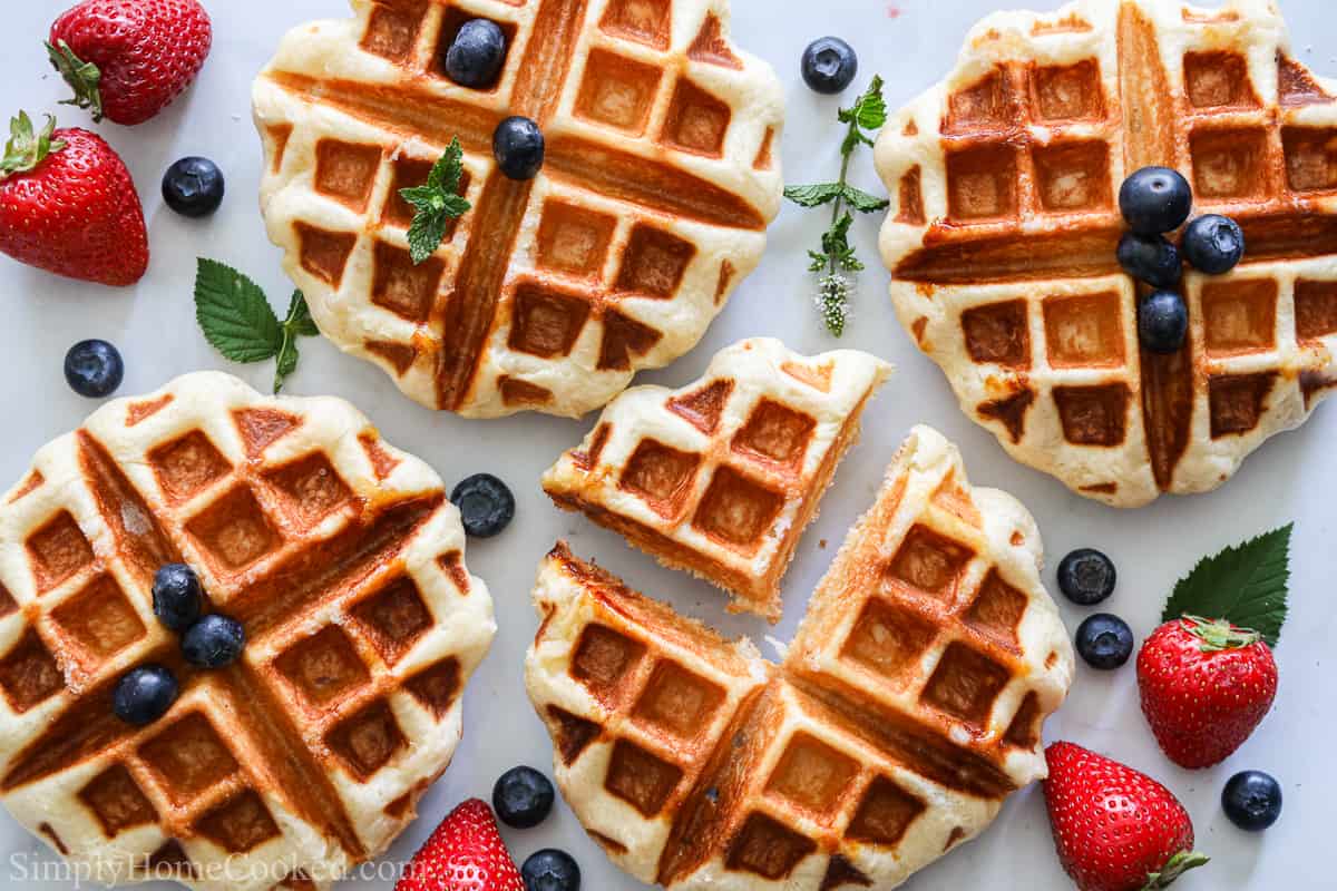 Buttery Belgian Liege Waffles scattered with blueberries, strawberries, and mint leaves on a white background.